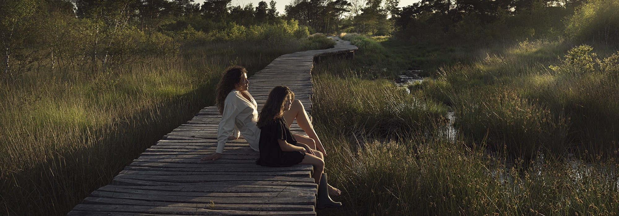 Moeder en dochter zitten op een houten knuppelpad in Nationaal Park De Groote Peel, omringd door moeras en riet in zacht avondlicht.