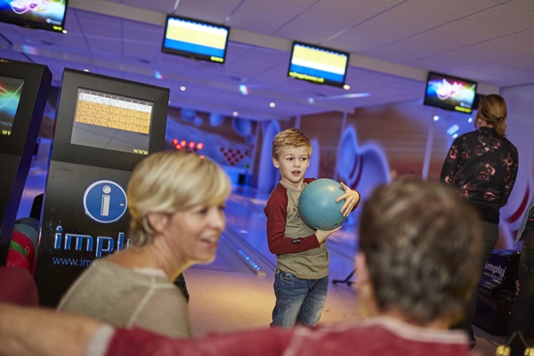 Ein Junge hält eine Bowlingkugel, während er sich zum Wurf bereit macht, während seine Eltern im Vordergrund beim Hyperbowling im Freizeitpark de Leistert miteinander reden.