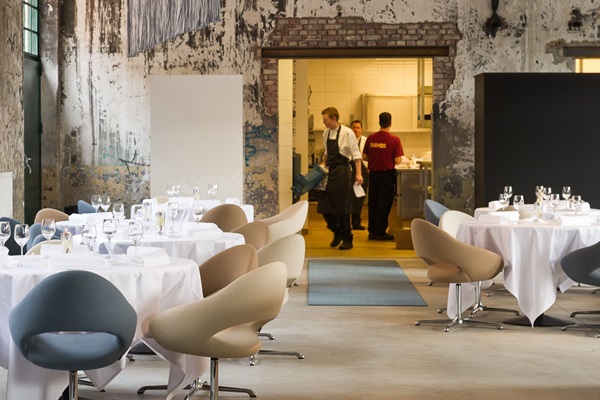 Die Köche des Restaurant One sind in der Küche im Hintergrund beschäftigt, während die Stühle und Tische für die Gäste in einer industriellen Umgebung eingedeckt werden