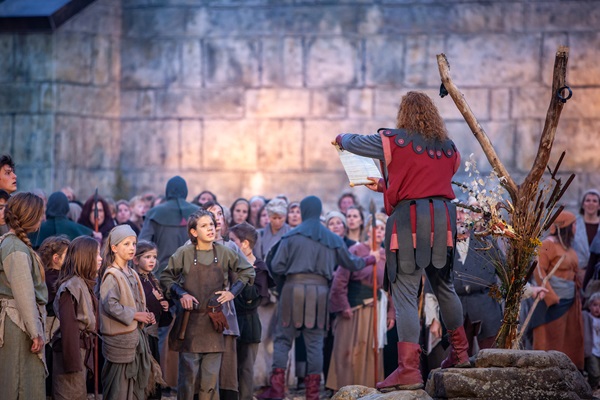 Middeleeuws theaterspektakel met een grote groep acteurs in historische kostuums op een podium voor een stenen muur. Een man met een rood-zwart kostuum leest een perkament voor aan het publiek.