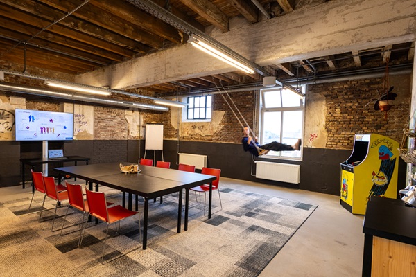 Industriële ruimte met houten balken, bakstenen muren, een vergadertafel met rode stoelen en een persoon op een schommel bij het raam.