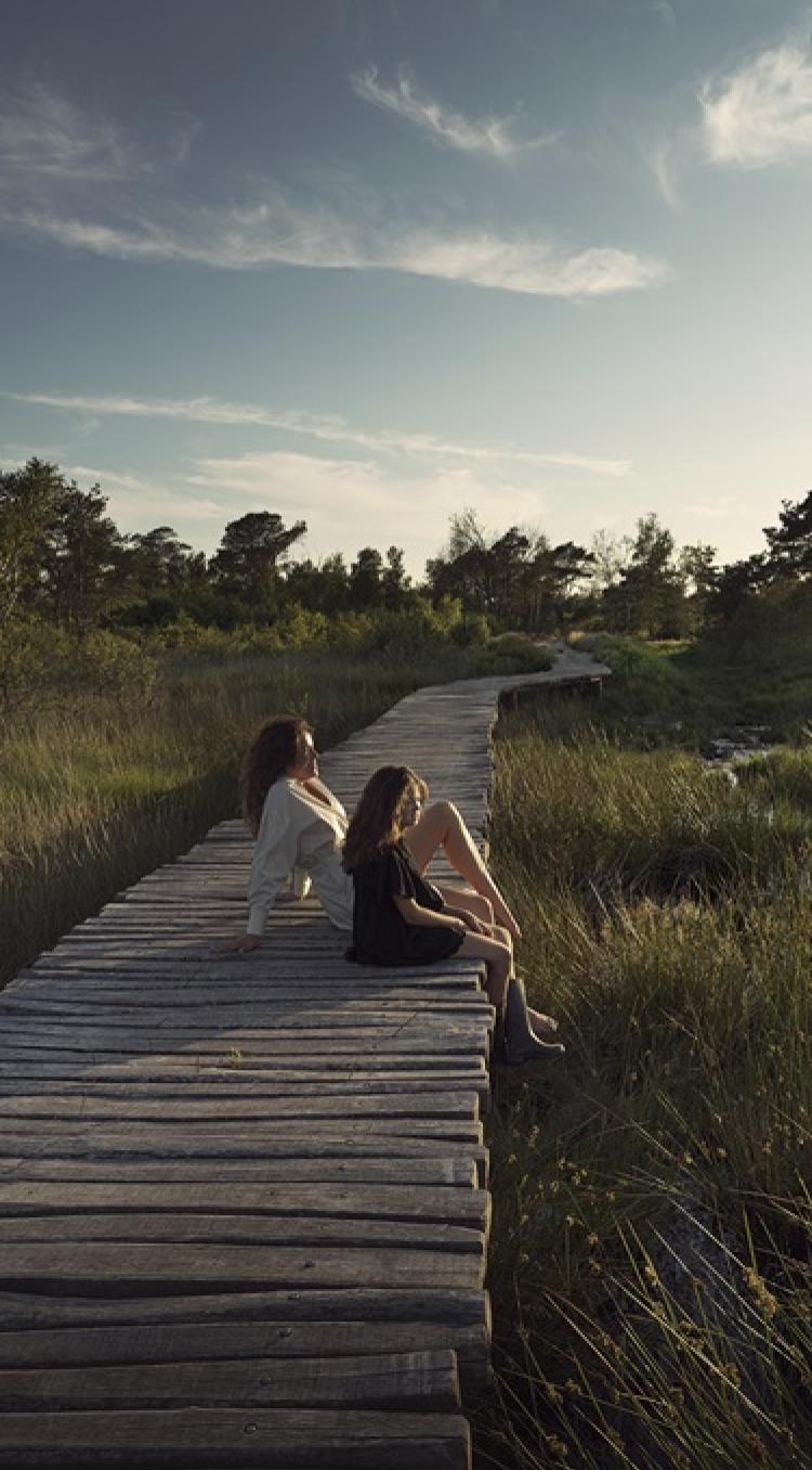 Moeder en dochter zitten op een houten knuppelpad in Nationaal Park De Groote Peel, omringd door moeras en riet in zacht avondlicht.