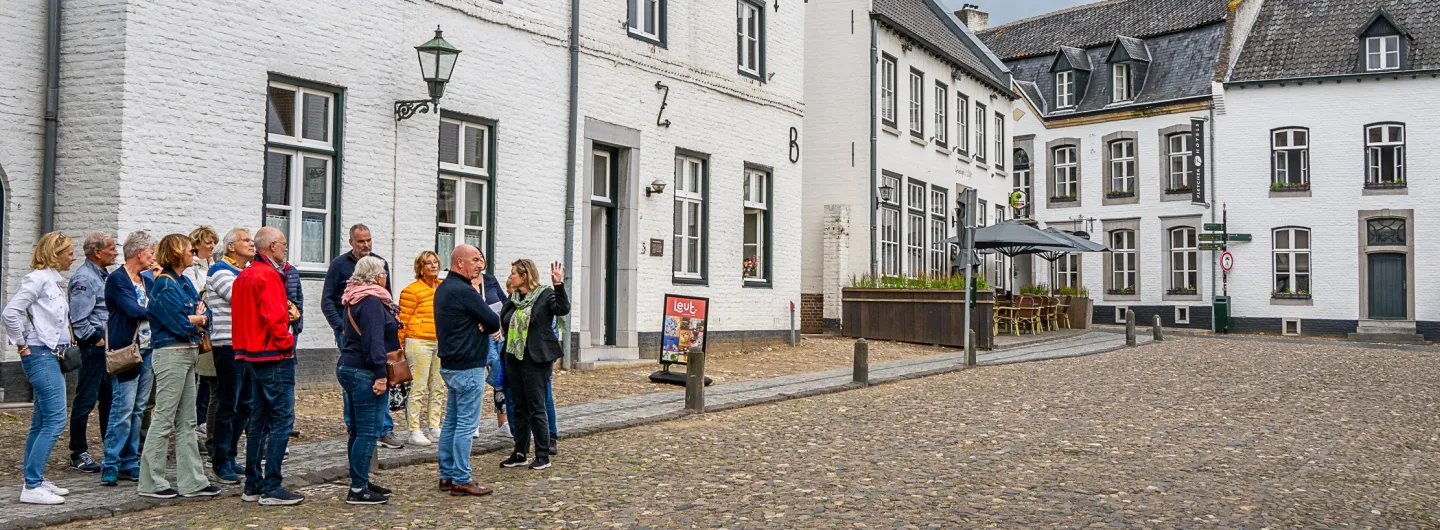 Een groep mensen volgt een rondleiding door een sfeervol historisch dorp met witgekalkte huizen, geplaveide straten en klassieke lantaarnpalen. De gids vertelt enthousiast terwijl de groep aandachtig luistert.