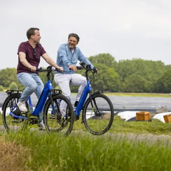 Twee mannen fietsen op blauwe e-bikes langs aspergevelden, omringd door groen en een open landschap. Ze lachen en kijken naar elkaar terwijl ze ontspannen fietsen.