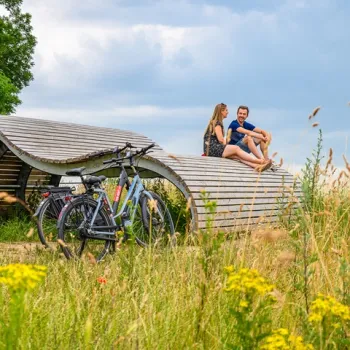 Twee fietsen geparkeerd bij een moderne houten rustplek, waar een stel geniet van het uitzicht tussen wilde bloemen en gras.