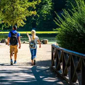 Twee wandelaars op een pad langs het water, met rugzakken en zomerse kleding, omringd door groen en zonlicht.
