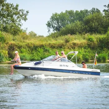Vier personen varen met een motorboot over de Maasplassen, met zomerse kleding en groene oever op de achtergrond.