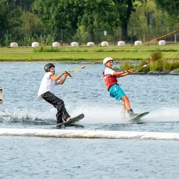 Twee jongeren doen aan waterskiën met een kabelbaan op een recreatieplas, met grasland en bomen op de achtergrond.