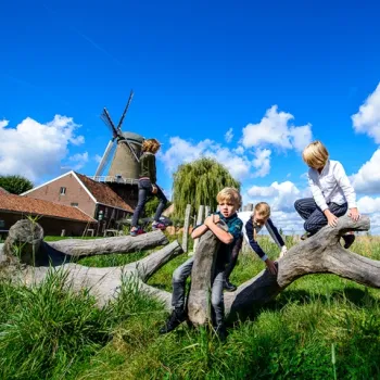 Kinderen klimmen op boomstammen in speeltuin OERRR in Stevensweert, met de Hompesche Molen en een bijgebouw van de molen op de achtergrond.