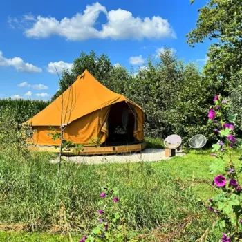 Geelbruine glampingtent tussen het groen in een voedselbos bij Eindje Buiten, omringd door bloemen, bomen en zomerse rust.