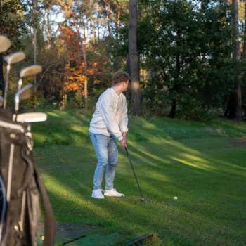 Golfer op de groene par 3-baan van De Herkenbosche, met golfclubs op de voorgrond en bos op de achtergrond.