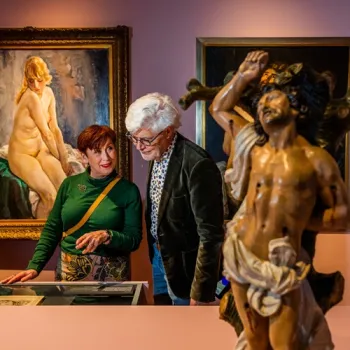 Twee bezoekers bekijken samen een museumvitrine, omringd door schilderijen en een groot religieus beeld.